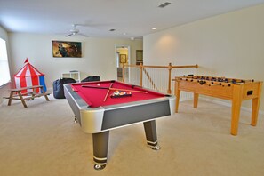 Une table de billard recouverte d’un feutre rouge, une table de baby-foot et une petite tente de jeu dans une pièce dont le sol est recouvert de moquette et qui est équipée d’un ventilateur de plafond.