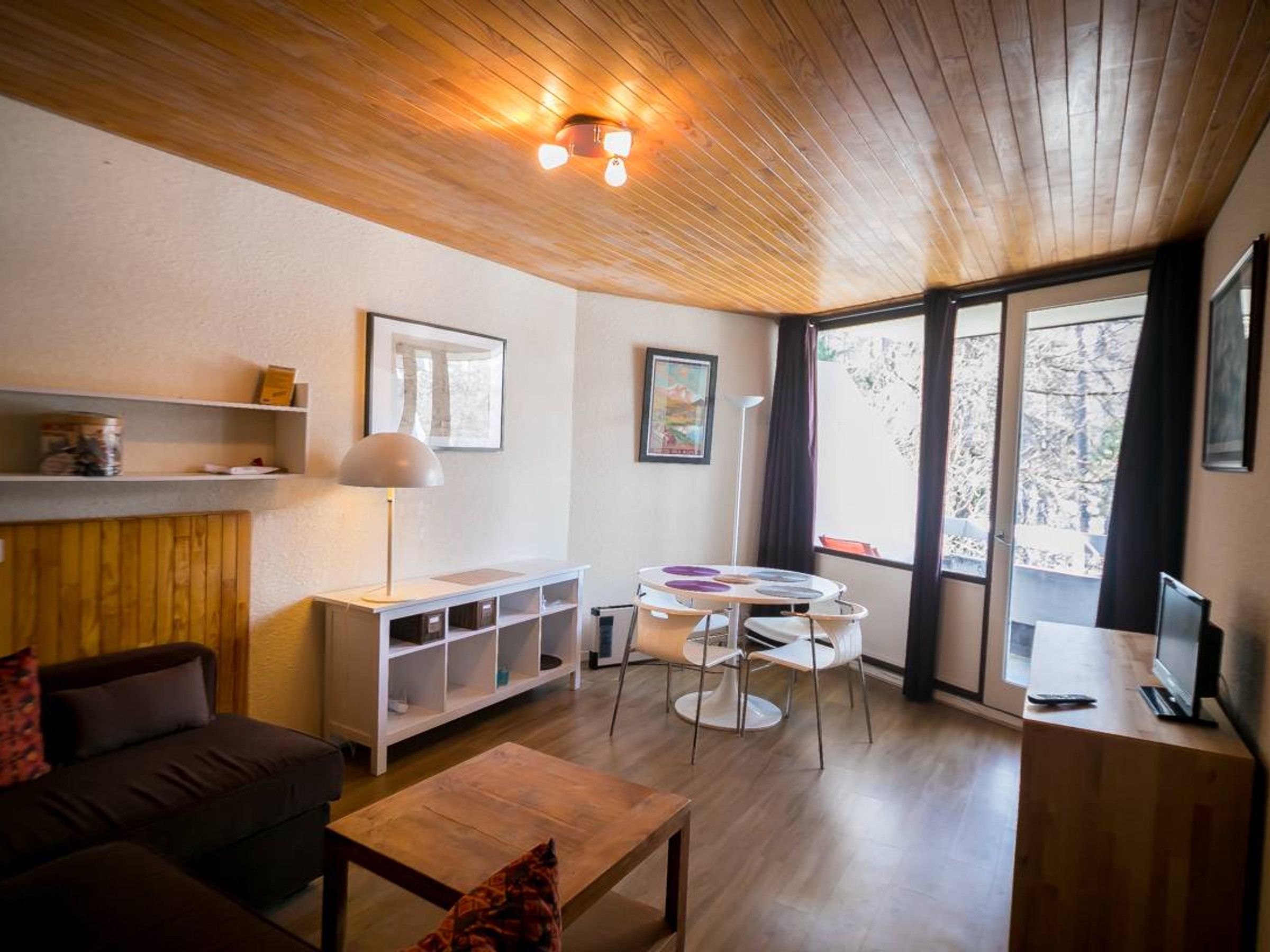 A modern living room with a wooden ceiling, a sofa, a coffee table, a TV stand, and a dining area with chairs.