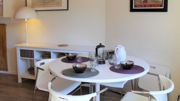 Un coin repas avec une table ronde dressée pour quatre personnes, entourée de chaises blanches. On y trouve un meuble blanc avec des étagères sous la table, ainsi qu’une carte encadrée et une affiche accrochées au mur.
