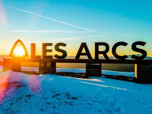 Eine Brücke, auf der in großen Buchstaben „ALLES ARCS“ steht, mit einer untergehenden oder aufgehenden Sonne im Hintergrund und Schnee auf dem Boden.