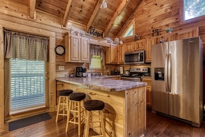 A cozy cabin-style kitchen with wooden walls, a large island with stools, stainless steel appliances, and a clock on the wall.