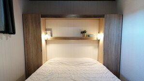 Ein schmaler Raum mit einem Bett, einem Holzkopfschild und einem Regal mit einer Pflanze und einer Lampe.