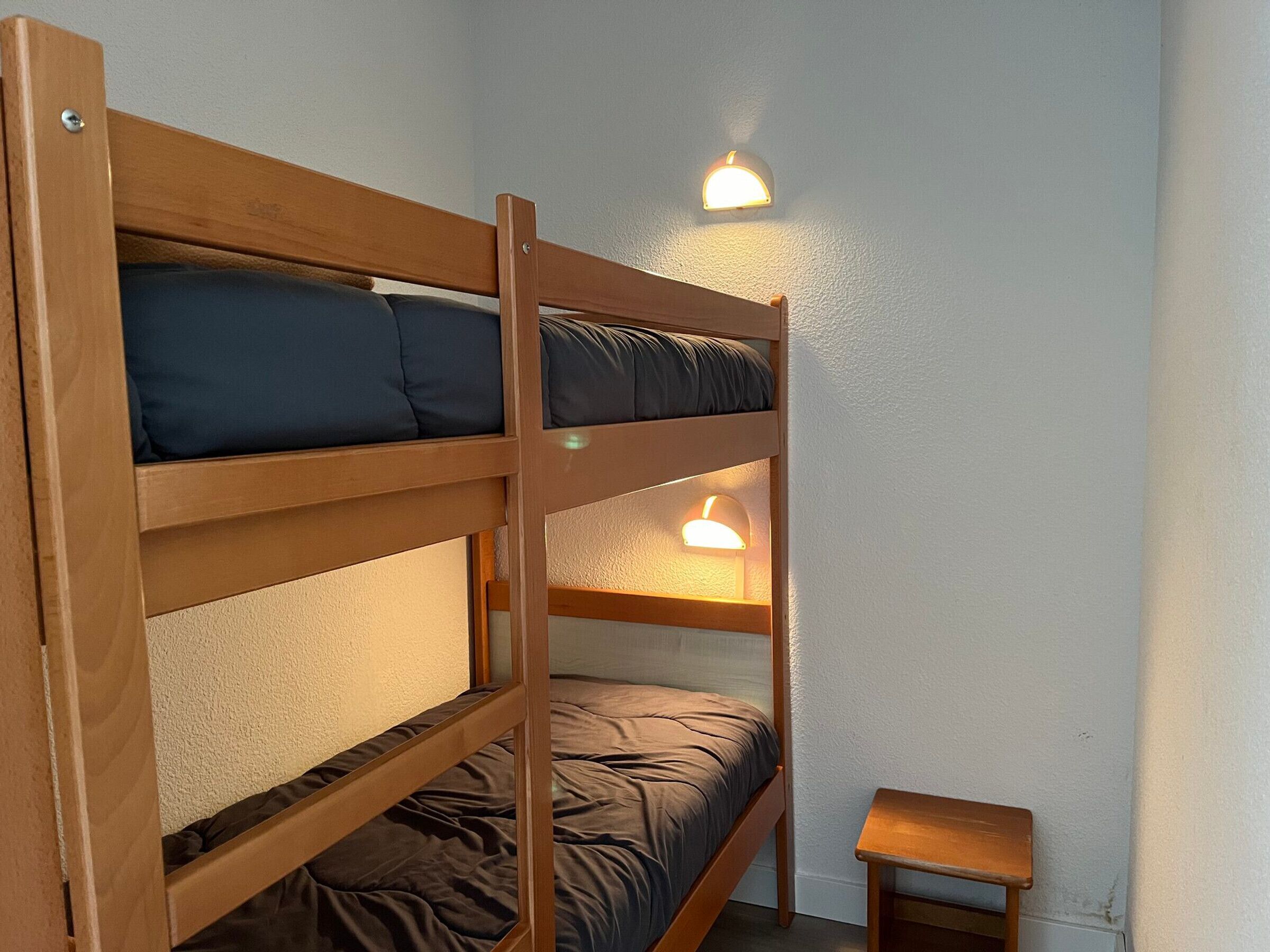 A bunk bed with two levels, each with a pillow and a blanket. There are two wall-mounted lights above the bed. A small wooden stool is placed beside the bed.