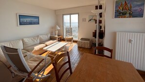 Ein Wohnzimmer mit einer Couch, einem Holztisch und einem Sessel. Ein Fenster bietet Blick auf die Stadt, und an der Wand hängt ein Gemälde.