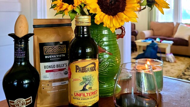 Une table avec une bouteille d’huile d’olive extra vierge, un paquet de café en grains de la marque Bongo Billy’s, une bouteille de Paesano et un vase rempli de tournesols et de feuilles vertes.