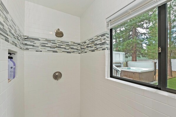 A bathroom with white tiles, a shower, a window with a view of a hot tub and trees.