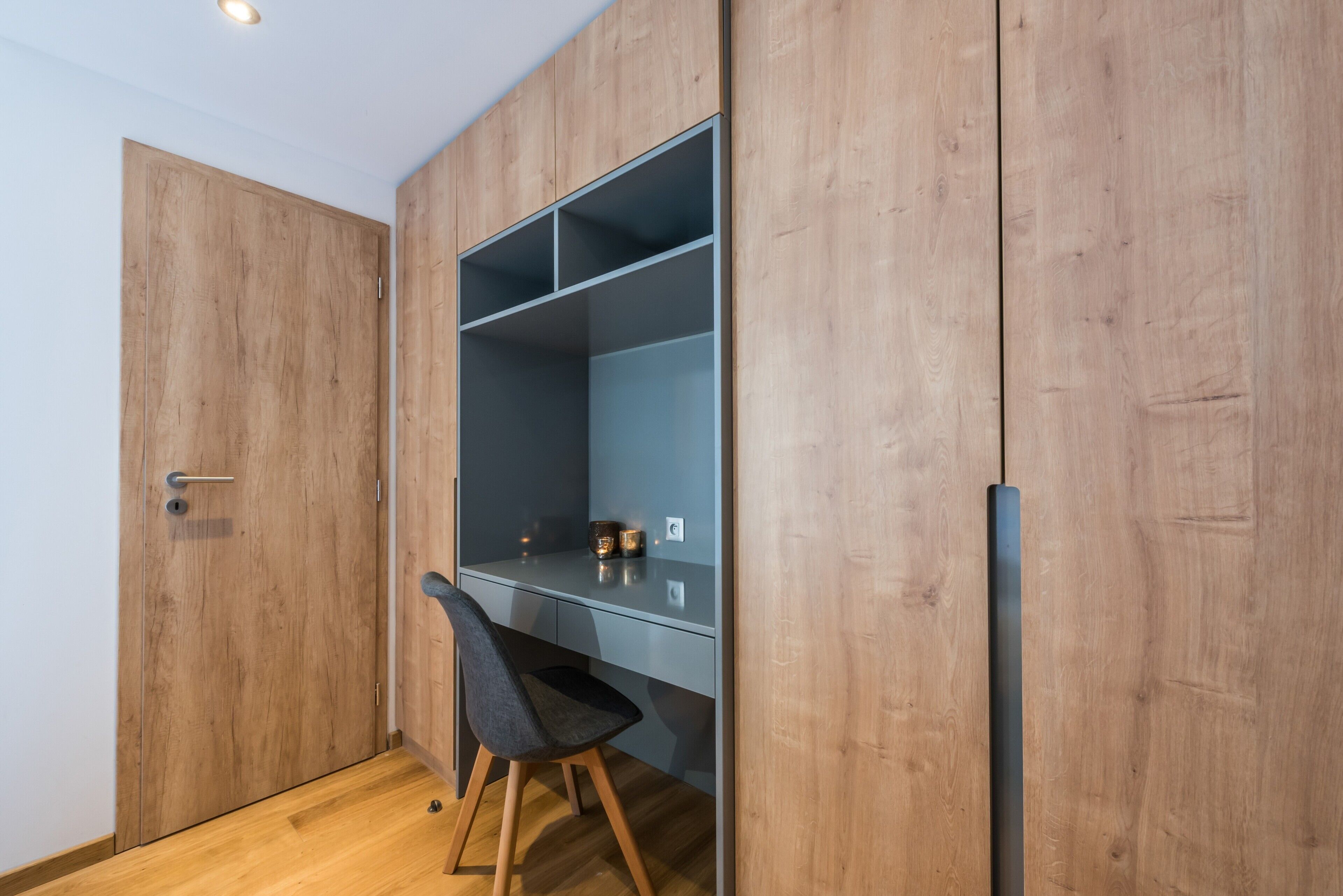 A room with a wooden wardrobe, a desk with a chair, and a shelf unit.