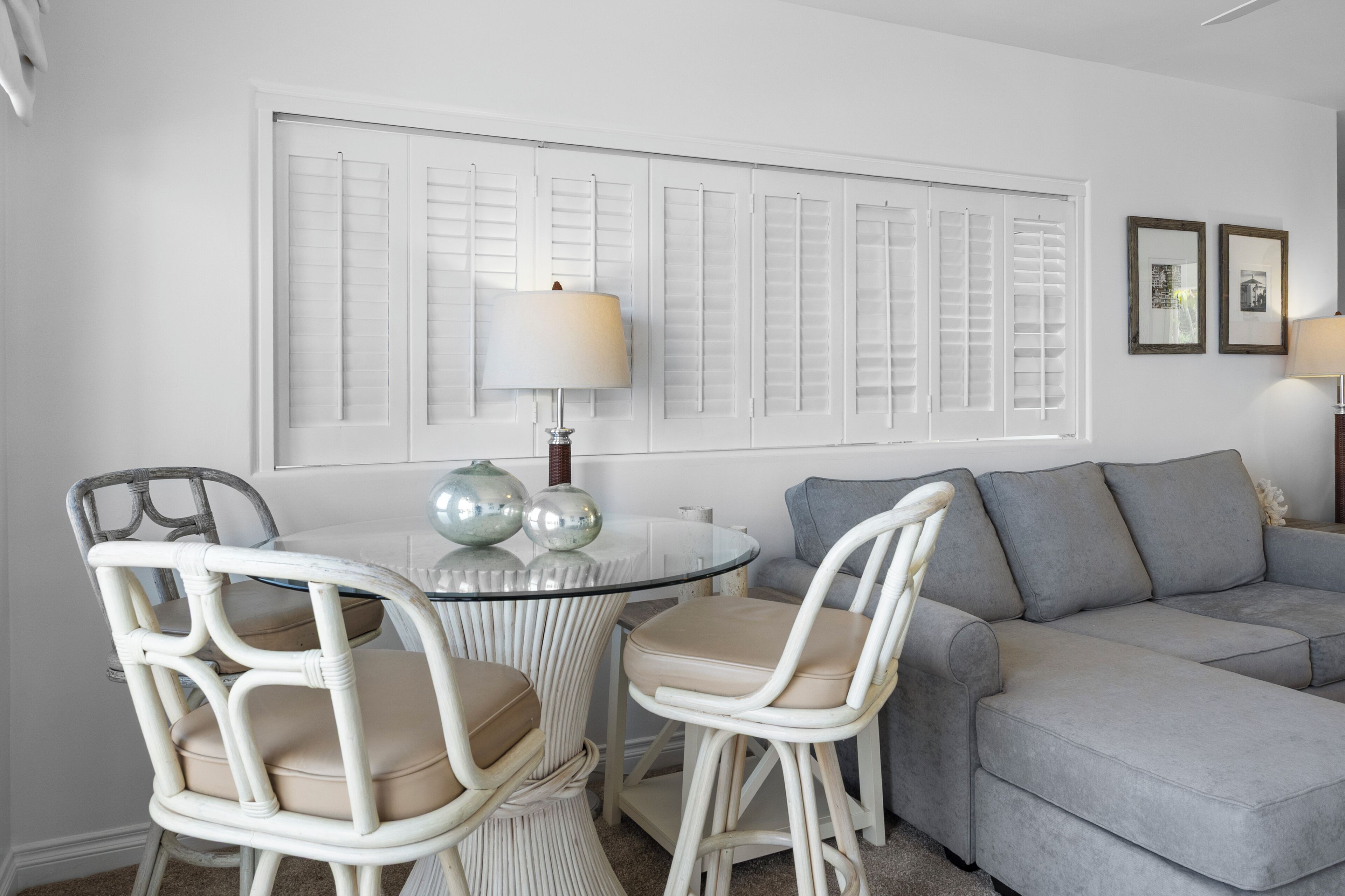 A living room with a grey sofa, two white wicker chairs, a glass table, and a window with shutters.