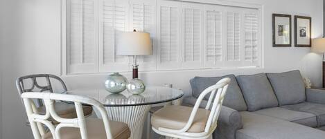 A living room with a grey sofa, two white wicker chairs, a glass table, and a window with shutters.
