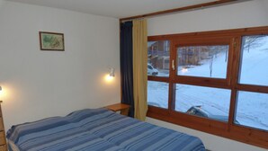 A bedroom with a bed, a nightstand, a window with a view of a snowy landscape, and a wall-mounted lamp.