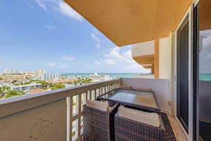 A balcony with wicker furniture, a view of the ocean, and city buildings in the background.