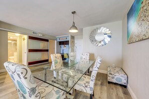 Un'area pranzo con un tavolo in vetro, sedie a fantasia, uno specchio decorativo a parete e un quadro a tema spiaggia.