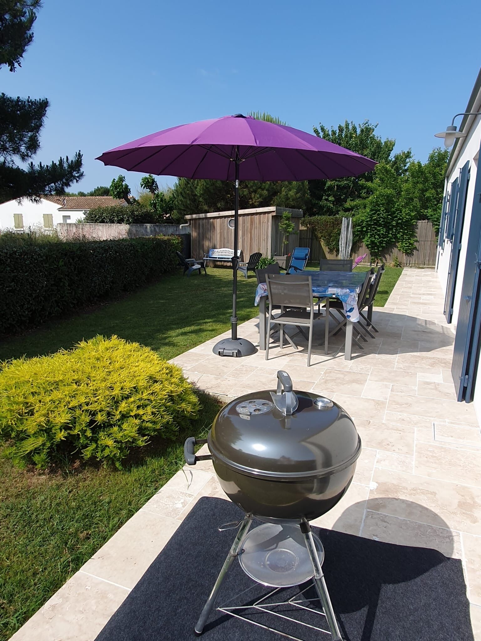 En grill i en baghave med et terrassemøbelset og stole under en lilla parasol.