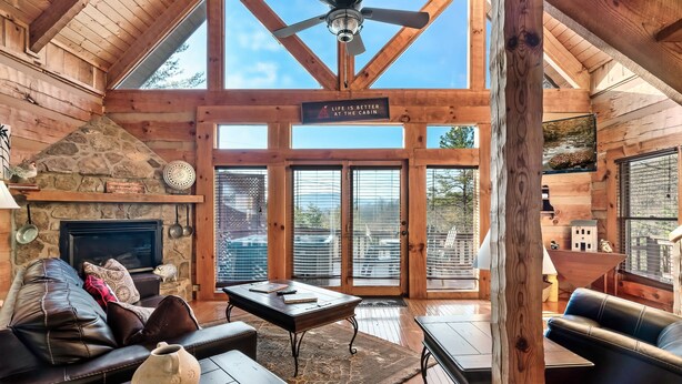 A cabin interior with a stone fireplace, leather furniture, and a large window overlooking a scenic view.