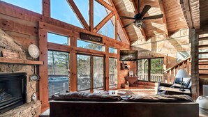 A cabin interior with a stone fireplace, leather sofa, wooden beams, and large windows.