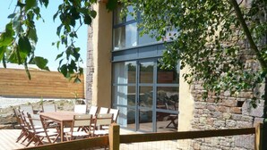 A patio with a wooden table and chairs, a stone wall, and a glass door leading to an interior space.
