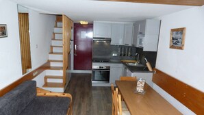 Eine kompakte Küche mit Kühlschrank, Backofen und Mikrowelle. Es ist ein Holzesstisch mit Stühlen und ein graues Sofa vorhanden. Eine Treppe führt auf eine obere Ebene.