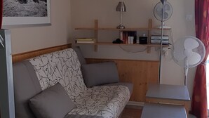 Et værelse med sofa, et lille bord, en ventilator og en reol med bøger og en lampe.