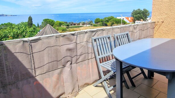 En balkon med et bord og stole, med udsigt over kystlandskab med grønt og bygninger.