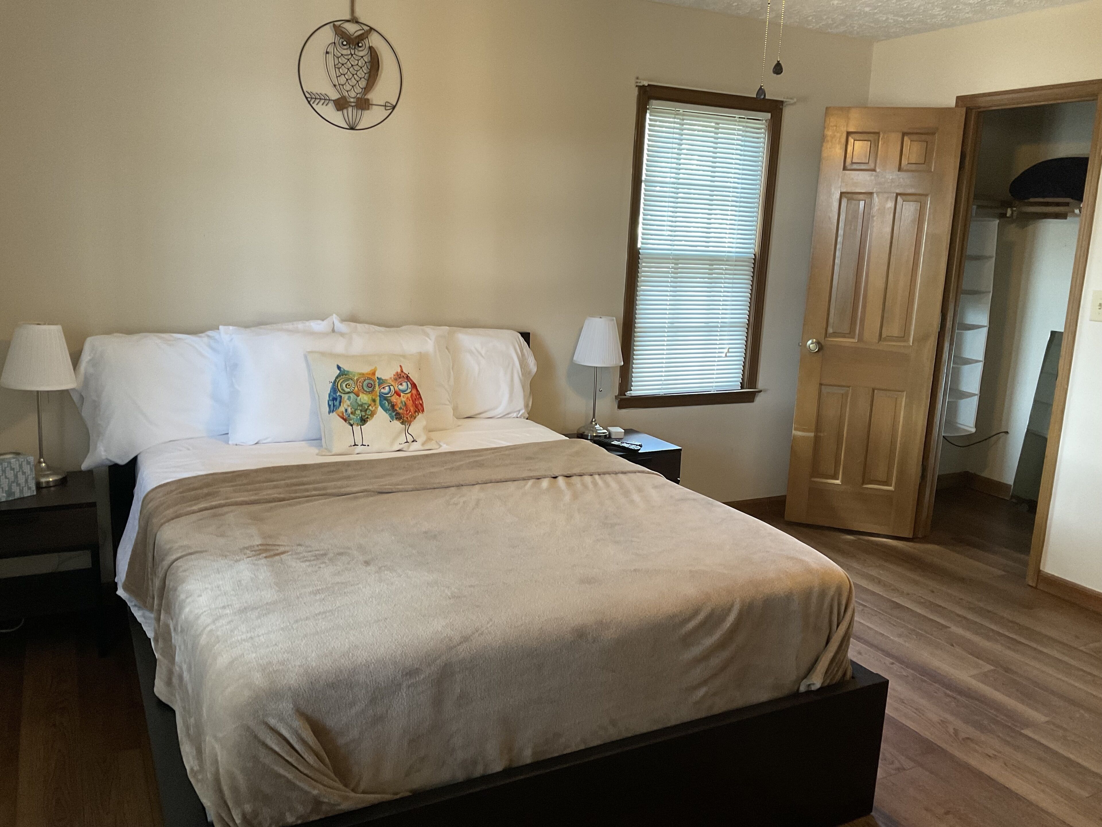 A bedroom with a bed, pillows, a nightstand, a lamp, a window with blinds, and a door.