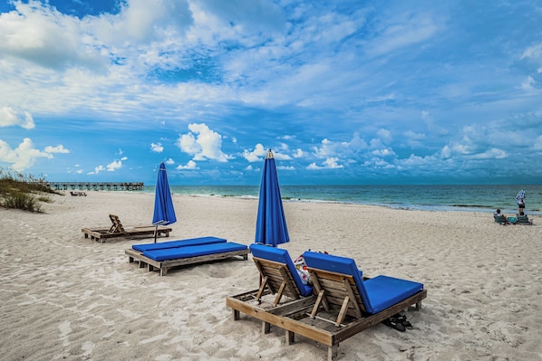 A beach with wooden lounge chairs and blue umbrellas, a pier in the background, and a clear sky.