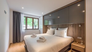 A modern bedroom with a large bed, a window with curtains, a bedside table with a lamp, and built-in cabinets. - Ipê - Modern | Sauna | Morzine (Montriond)