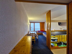A room with a wooden table, a bed with a view, and a kitchenette.