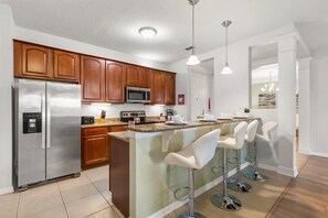 A modern kitchen with a stainless steel refrigerator, wooden cabinets, a microwave, and a bar area with stools.