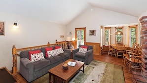A spacious living room with a high ceiling, a wooden dining table with chairs, a grey sofa with patterned cushions, and a wooden coffee table. - Big Tree Lodge by Destination Big Bear : Ping Pong! Wood Burning Fireplace! Cable! Internet! Propane BBQ! (Big Bear City)