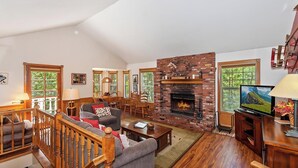 A spacious living room with a fireplace, wooden furniture, and large windows. - Big Tree Lodge by Destination Big Bear : Ping Pong! Wood Burning Fireplace! Cable! Internet! Propane BBQ! (Big Bear City)