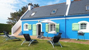 Una casa con fachada azul, techo gris y un pequeño porche con tres sillones negros y un paraguas blanco.