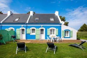 Una casa de dos pisos con fachada azul y blanca, una terraza de madera y un patio con mobiliario exterior.