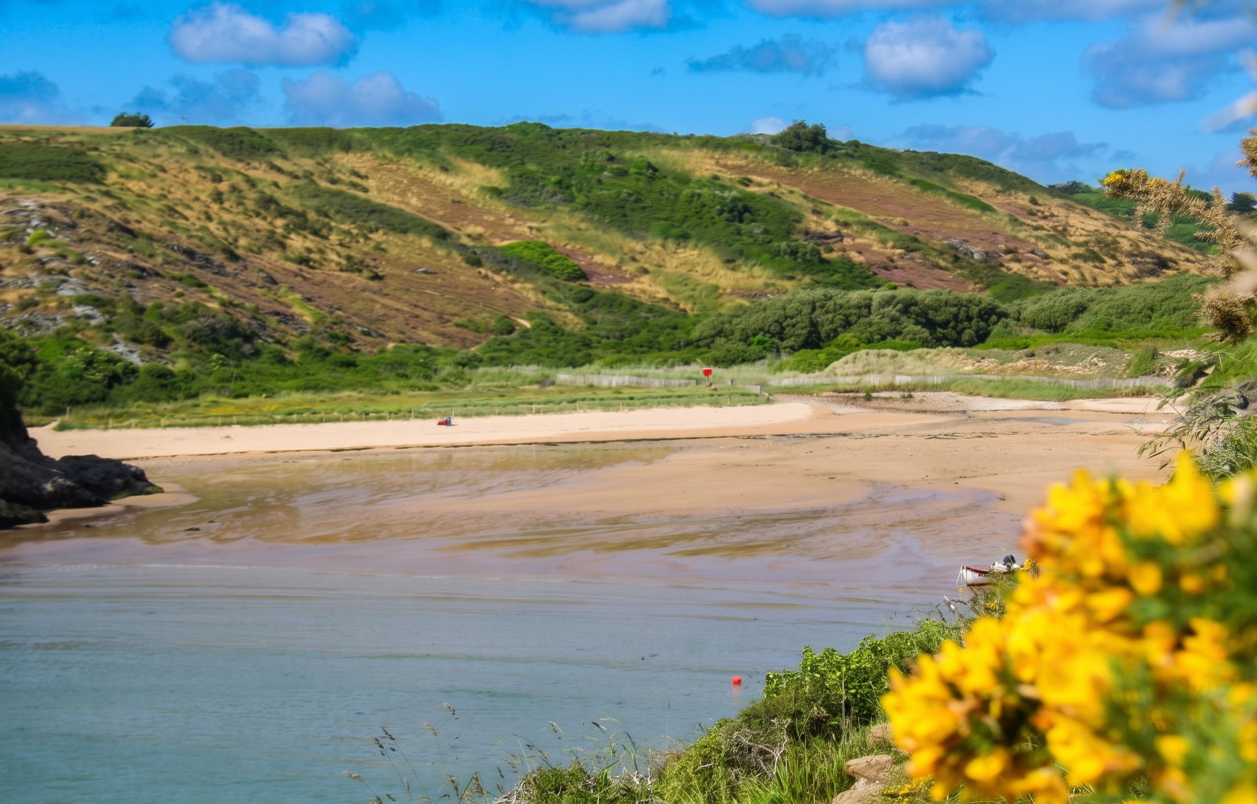 A coastal landscape with a sandy beach, calm water, and green hills in the background.