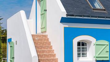 A two-story building with a blue exterior, a white roof, and a small window with green shutters.