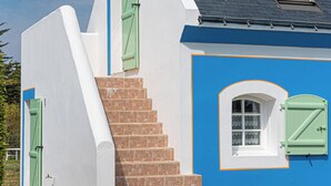 Un edificio con fachada azul, detalles blancos y contraventanas verdes. Cuenta con una escalera de ladrillo que conduce a una ventana con marco blanco.