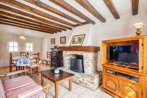A cozy living room with exposed wooden beams, a stone fireplace, a television, and a dining area with a table and chairs.