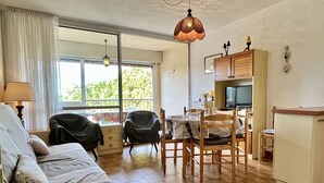 A cozy living room with a sofa, chairs, a dining table, and a TV. There is a large window providing a view of greenery outside. - 2-room apartment, beach access, parking, sleeps 4 - Argelès-sur-Mer (Argelès-sur-Mer)