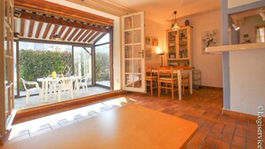 Una habitación con un amplio ventanal corredera que da a un espacio exterior, una mesa de comedor con sillas, una zona de cocina con armarios y una lámpara colgante.