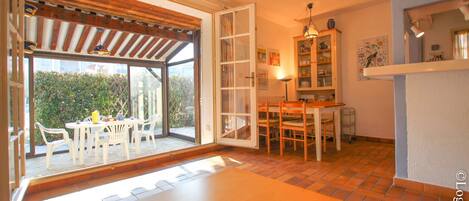 Une pièce dotée d’une grande porte-fenêtre coulissante donnant sur un espace extérieur, d’une table à manger avec des chaises, d’un coin cuisine avec des placards et d’un luminaire suspendu.