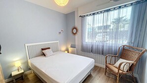 A bedroom with a bed, bedside lamps, a wicker chair, a window with curtains, and a ceiling fan. - Charming Renovated Apartment, Central Jullouville, 1 Bed, 4 Guests, Parking, 100m from Beach (Jullouville)