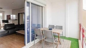 Un balcon avec une table et des chaises, une porte-fenêtre coulissante donnant sur le salon et un espace vert à l’extérieur.