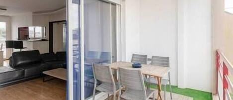 A balcony with a table and chairs, a sliding glass door leading to a living room, and a green area outside.