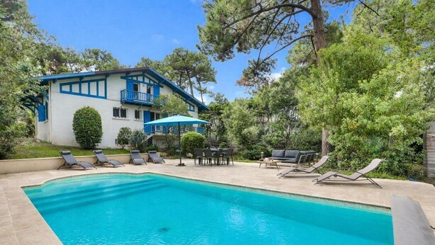 A pool area with lounge chairs, a dining table, and a building with a balcony. - Hossegor Lake, 10 pers Villa (SEIGNOSSE)
