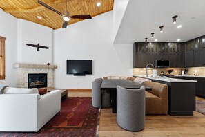 A modern living room with a stone fireplace, a flat-screen TV, a large sectional sofa, a dining table, and a kitchen area with dark cabinets and a countertop. - Fully Remodeled Townhome - Walk to Slopes, Pool, & Hot Tub! (Keystone)