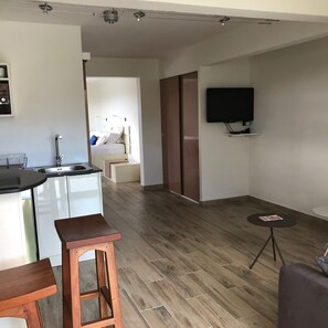 Cozy living space with a kitchen, dining, and seating area, featuring a flat-screen TV and a door to another room.