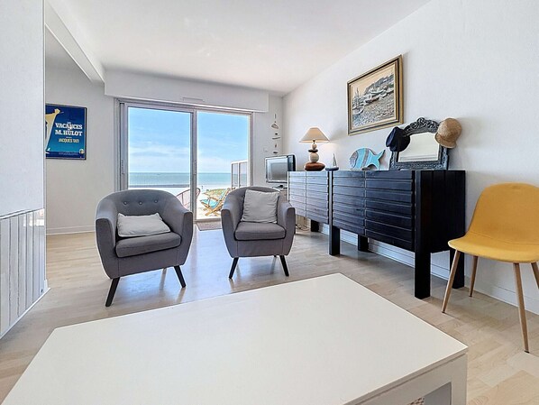 A modern living room with two grey armchairs, a white coffee table, a black dresser, and a yellow chair. A large window offers a view of the beach.