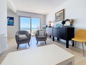 Uma sala de estar moderna com duas poltronas cinzentas, uma mesa de centro branca, um aparador preto e uma cadeira amarela. Uma grande janela oferece vista para a praia.