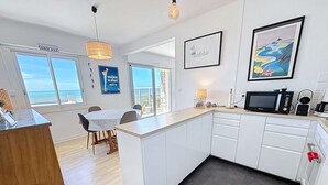 A modern kitchen with white cabinets, a wooden dining table, and a view of the sea through large windows. - Seaside apartment with terrace and parking (Jullouville)