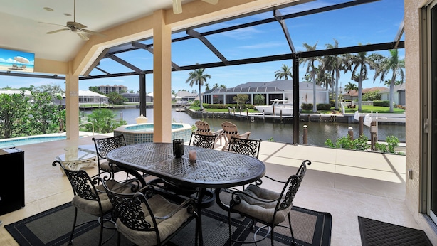 A covered outdoor area with a table and chairs, a ceiling fan, and a view of a water body and palm trees.