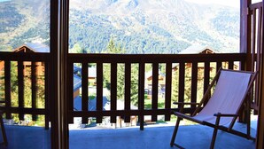 A balcony with a wooden railing, a single lounge chair, and a view of a mountainous landscape with buildings in the distance.